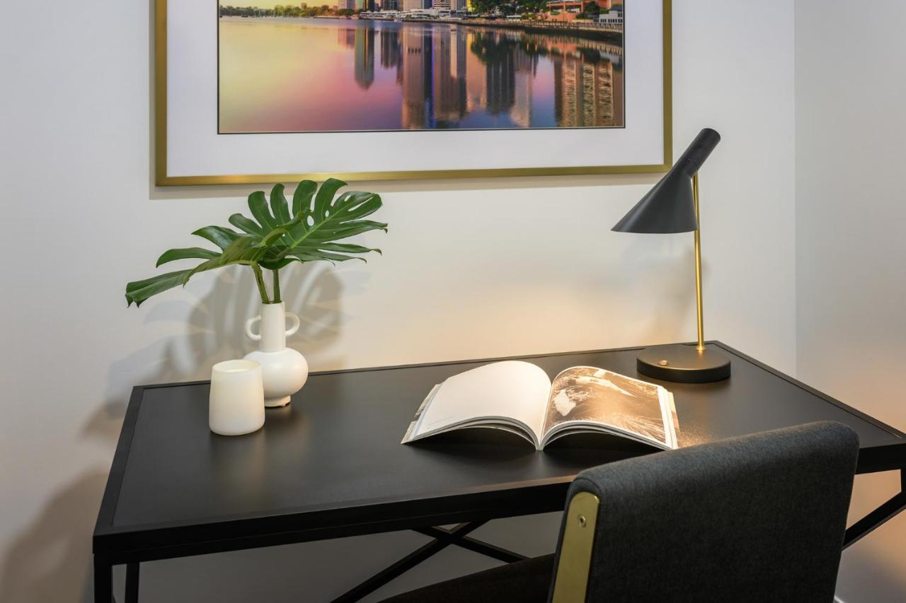 Meriton Suites Herschel Street, Brisbane Dış mekan fotoğraf The image shows a modern workspace featuring a black desk with a small open book on it. A stylish desk lamp provides lighting, and there is a minimalist white vase with a green plant beside it. There are also two small candles on the desk. Above the 