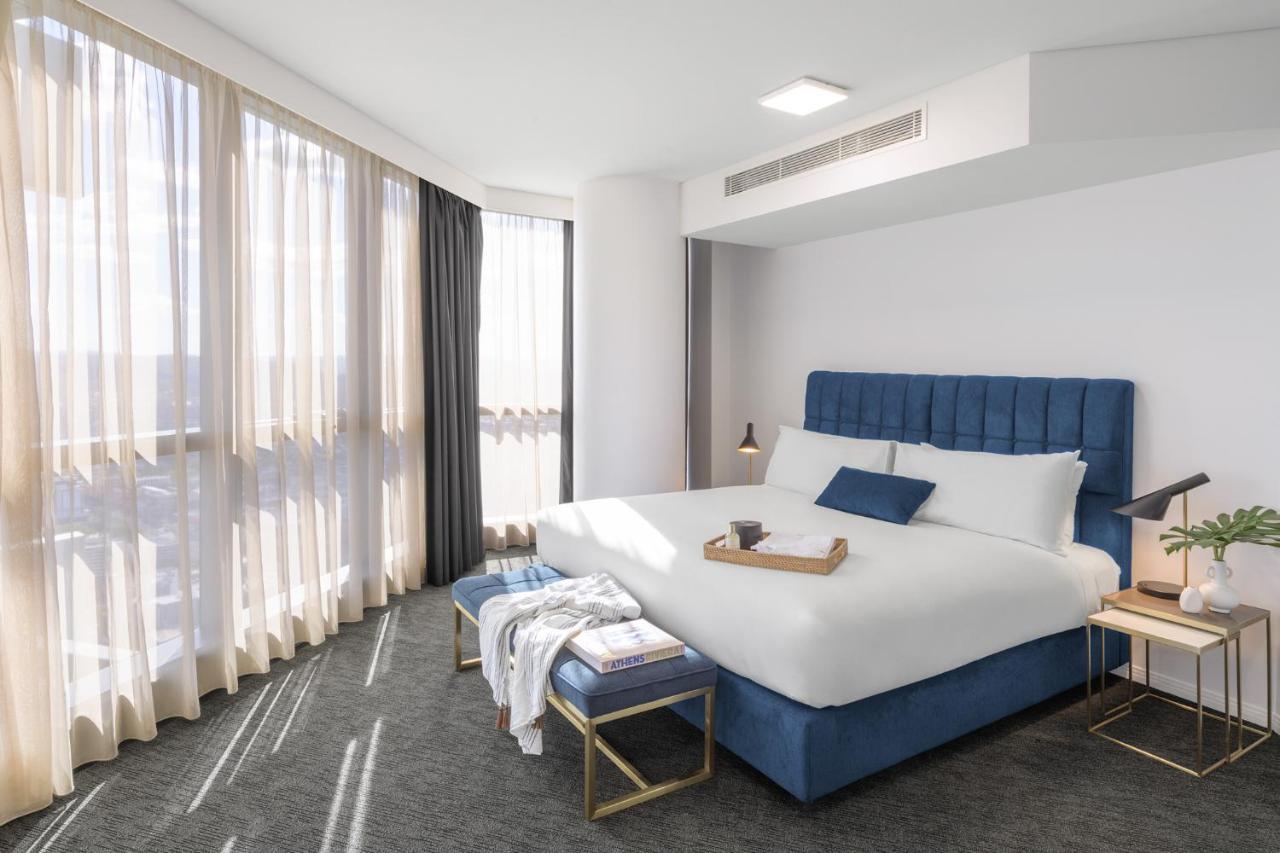 Meriton Suites Herschel Street, Brisbane Dış mekan fotoğraf The photo shows a modern hotel room featuring a large bed with a blue upholstered headboard and white bedding. There is a small tray on the bed, adding a decorative touch. Next to the bed, there is a blue bench and a bedside table with a lamp. The ro