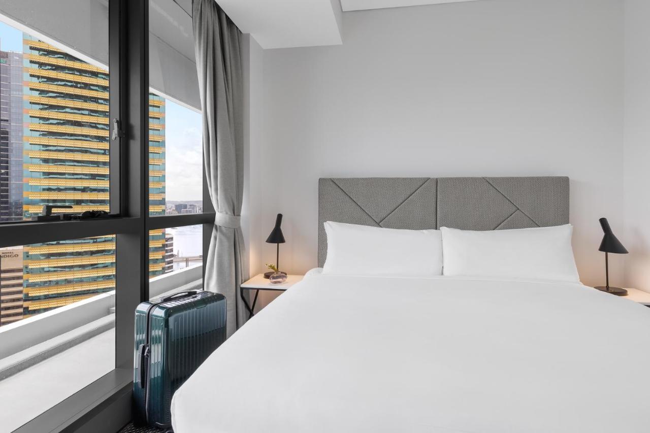 Meriton Suites Herschel Street, Brisbane Dış mekan fotoğraf The photo shows a modern hotel bedroom. It features a large bed with white bedding and two pillows. Behind the bed is a padded headboard in a neutral color. On either side of the bed, there are small bedside tables with lamps. A stylish piece of lugg