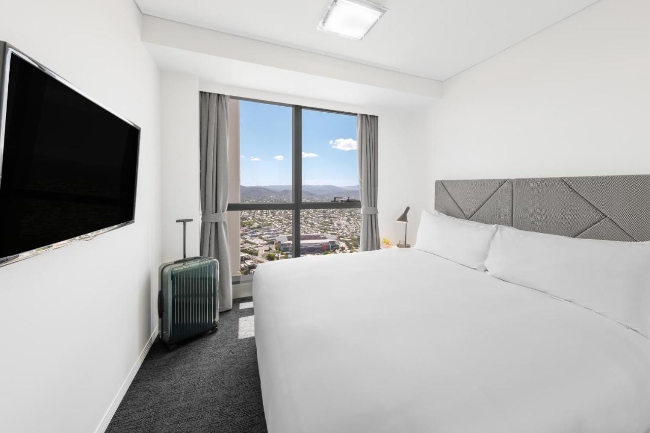 Meriton Suites Herschel Street, Brisbane Dış mekan fotoğraf The photo shows a modern hotel room. It features a large bed with white bedding and a gray headboard. There's a flat-screen TV mounted on the wall across from the bed. To the side, there's a small luggage trolley with a suitcase. The room has large w
