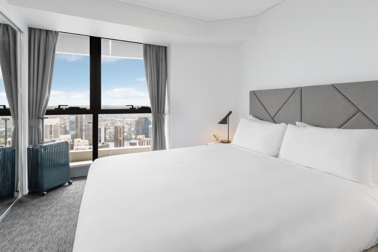 Meriton Suites Herschel Street, Brisbane Dış mekan fotoğraf The photo depicts a modern hotel bedroom featuring a large, neatly made bed with white linens. The room has a minimalist design with grey accents, including a geometric-patterned headboard. To the side of the bed, there is a small lamp and a blue sui