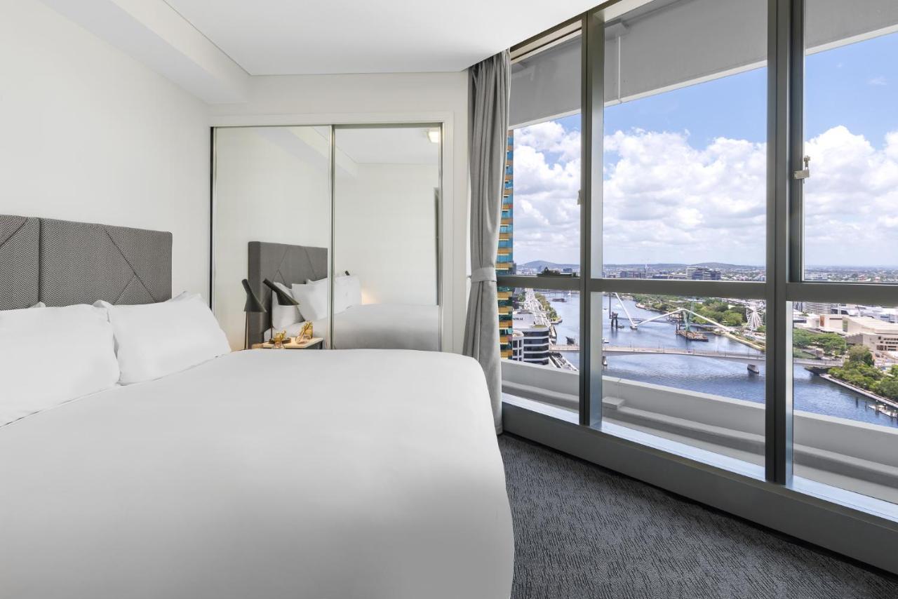 Meriton Suites Herschel Street, Brisbane Dış mekan fotoğraf The image shows a modern hotel room featuring a large bed with white bedding positioned against a wall. There is a large window providing a view of a river and the surrounding cityscape, with natural light streaming in. The room is furnished with a m